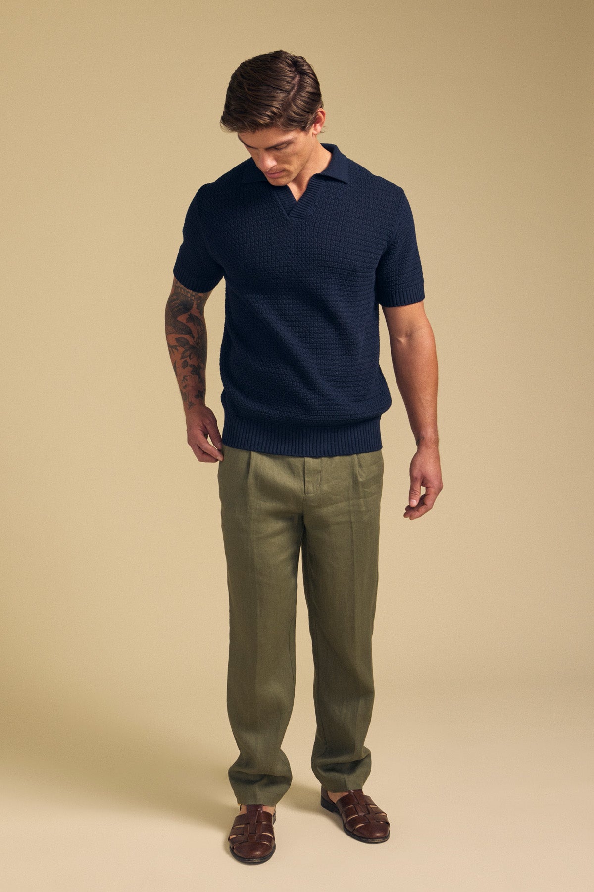 A man looks down, wearing the CARSTON SWEATER POLO by Velvet by Graham & Spencer in navy, paired with olive trousers and brown sandals against a plain beige background.