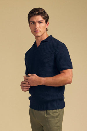 A man with brown hair wears the Velvet by Graham & Spencer CARSTON SWEATER POLO in navy with olive pants, standing against a neutral beige background.