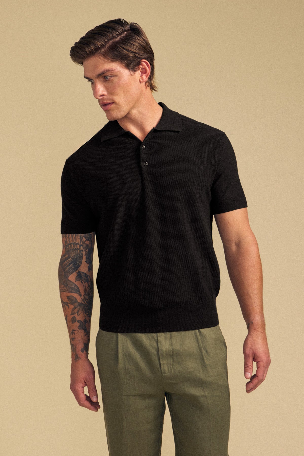 A man wearing the DILLON SWEATER POLO by Velvet by Graham & Spencer in black, paired with olive green pants, stands against a plain beige background, looking to his left.-41759794921665