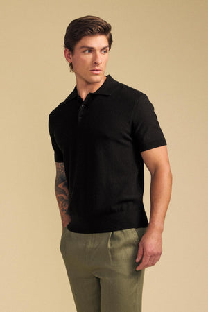 A man wearing the DILLON SWEATER POLO by Velvet by Graham & Spencer—a black textured knit short-sleeve polo shirt—paired with light olive-green pants stands against a plain beige background, looking to his left.