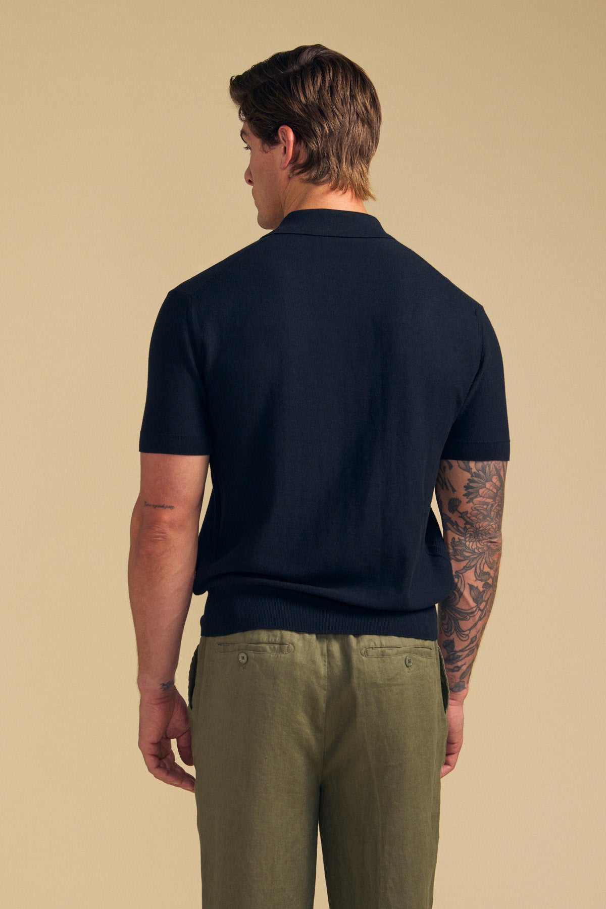 A man stands with his back to the camera, showcasing his tattooed right arm while wearing the DILLON SWEATER POLO— a black short-sleeve cotton polo from Velvet by Graham & Spencer—paired with olive green pants, set against a plain beige background.
