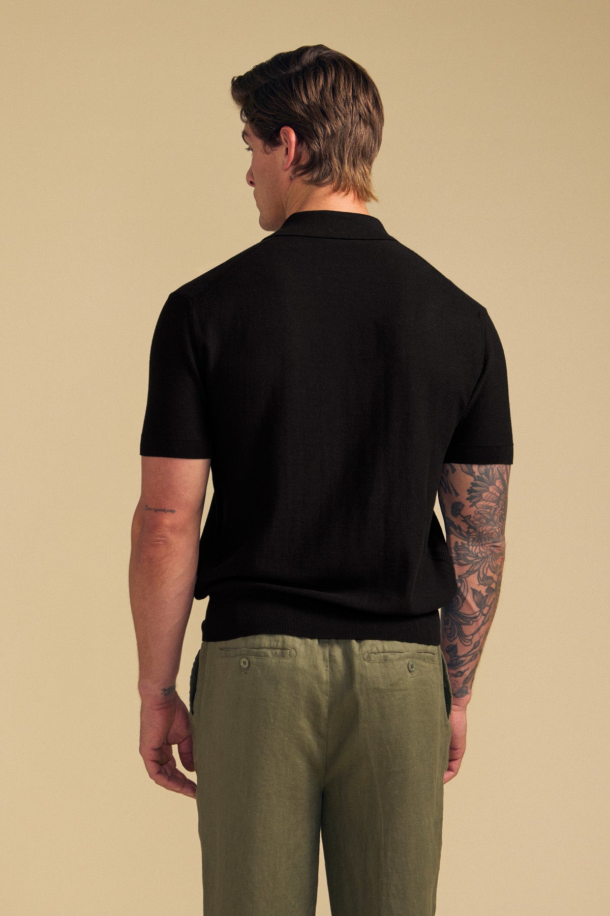 A man stands with his back to the camera, showcasing his tattooed right arm while wearing the DILLON SWEATER POLO— a black short-sleeve cotton polo from Velvet by Graham & Spencer—paired with olive green pants, set against a plain beige background.-41759794823361