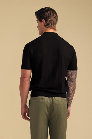 A man stands with his back to the camera, showcasing his tattooed right arm while wearing the DILLON SWEATER POLO— a black short-sleeve cotton polo from Velvet by Graham & Spencer—paired with olive green pants, set against a plain beige background.