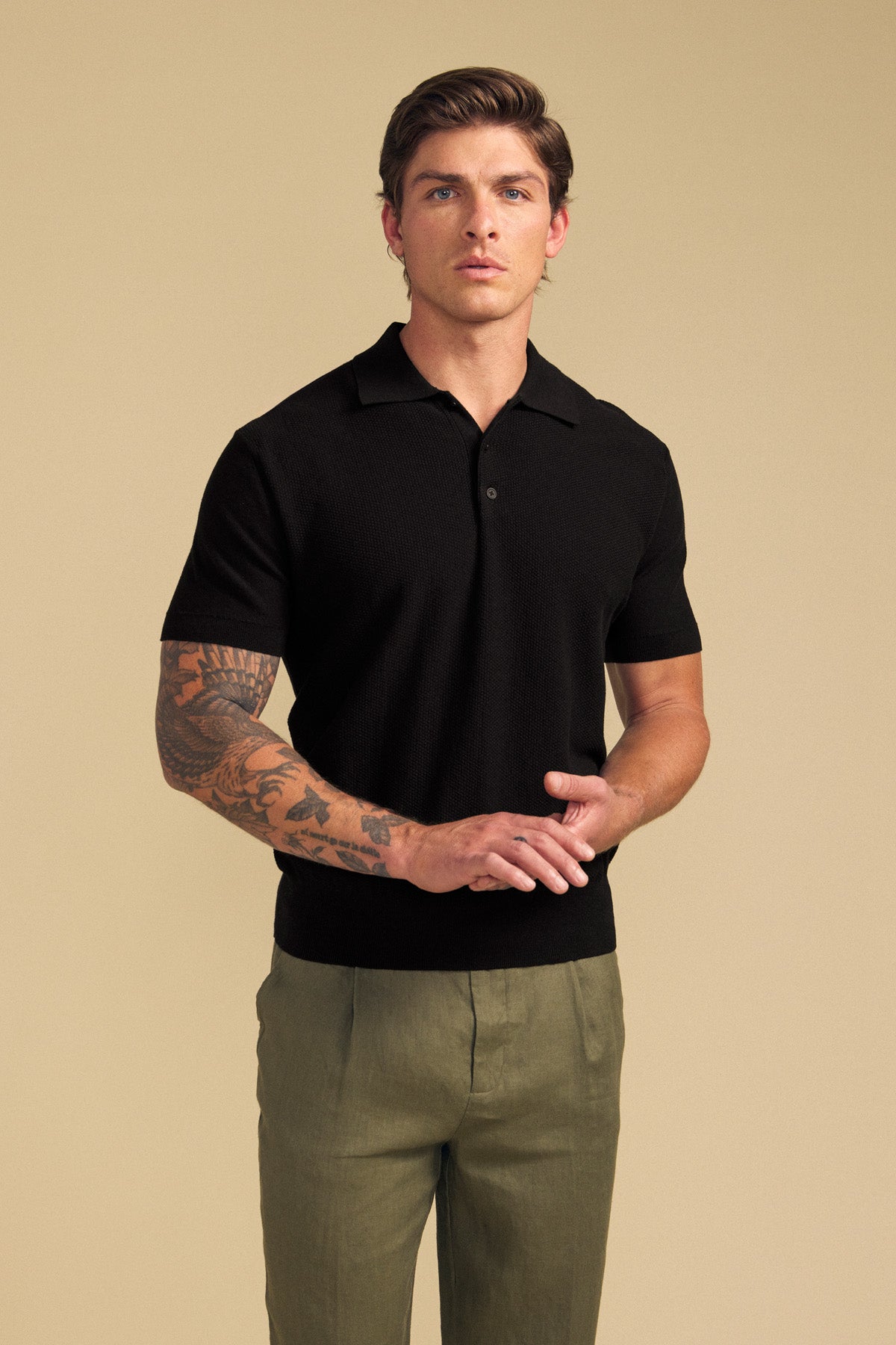 A man stands against a beige background wearing the DILLON SWEATER POLO in black by Velvet by Graham & Spencer—this short-sleeve cotton sweater is paired with olive green pants, offering a stylish warm-weather layer, while his arm tattoos remain visible.
