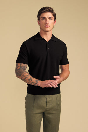 A man stands against a beige background wearing the DILLON SWEATER POLO in black by Velvet by Graham & Spencer—this short-sleeve cotton sweater is paired with olive green pants, offering a stylish warm-weather layer, while his arm tattoos remain visible.