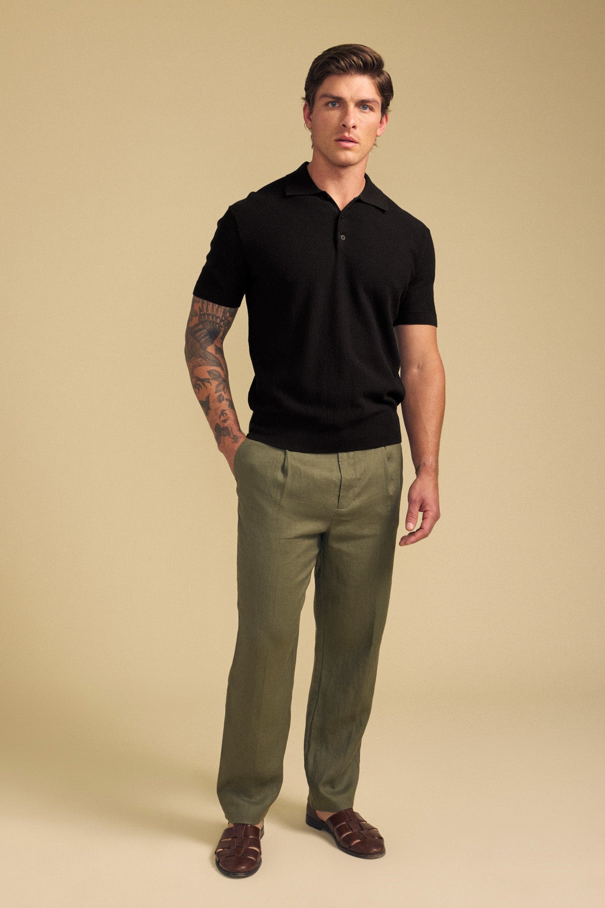 A man stands against a beige background wearing the Velvet by Graham & Spencer DILLON SWEATER POLO—a black textured knit short-sleeve shirt—paired with olive green trousers, brown shoes, and showcasing a tattoo on his right arm.