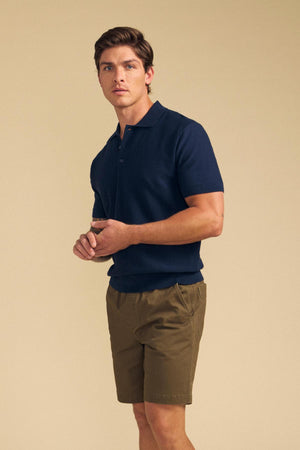 A man stands against a beige background wearing the Velvet by Graham & Spencer DILLON SWEATER POLO in navy with olive green shorts—a perfect warm-weather layer.