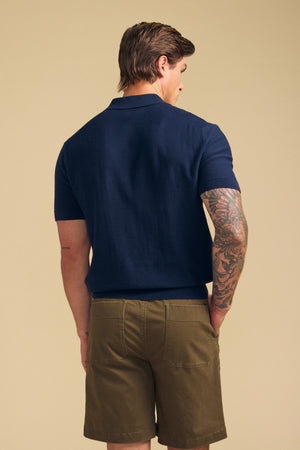 A man with a tattooed right arm wears the navy DILLON SWEATER POLO by Velvet by Graham & Spencer as a lightweight layer, paired with olive shorts. He faces away from the camera against a plain beige background.