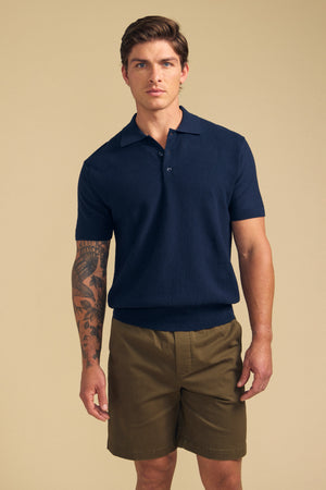 A man in the DILLON SWEATER POLO by Velvet by Graham & Spencer and olive shorts stands against a beige background, showing his left forearm tattoo and short brown hair—ready for any warm-weather layering.