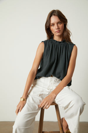 A woman is sitting on a stool in white pants and a black BIANCA COTTON GAUZE TANK TOP by Velvet by Graham & Spencer.