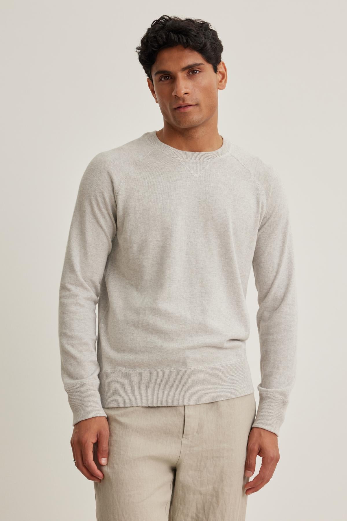   A person stands against an off-white background, wearing the DARIO COTTON CASHMERE SWEATER by Velvet by Graham & Spencer and beige pants, looking at the camera. 