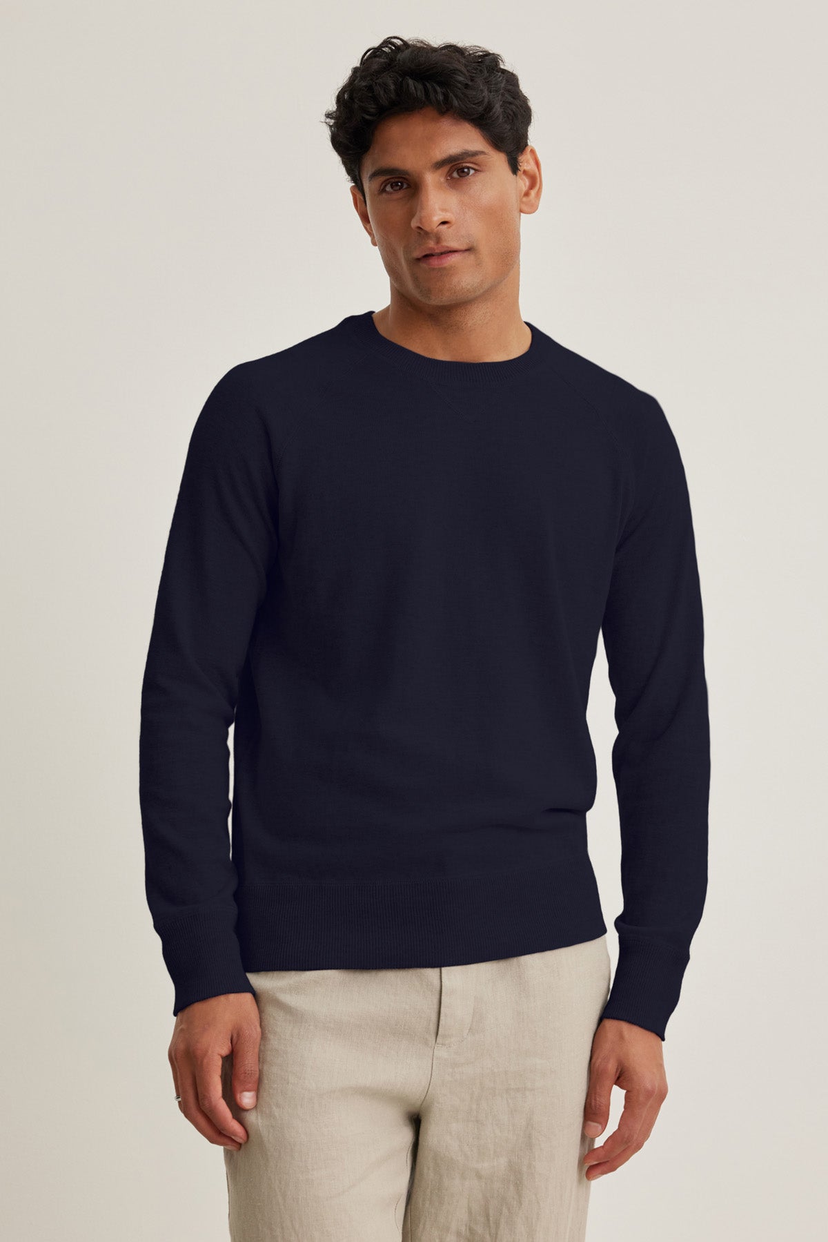 A man wearing the DARIO SWEATER by Velvet by Graham & Spencer—a plain dark navy mid-weight cotton cashmere crewneck—paired with light beige pants, stands against a neutral background facing the camera with a neutral expression.-41008986816705