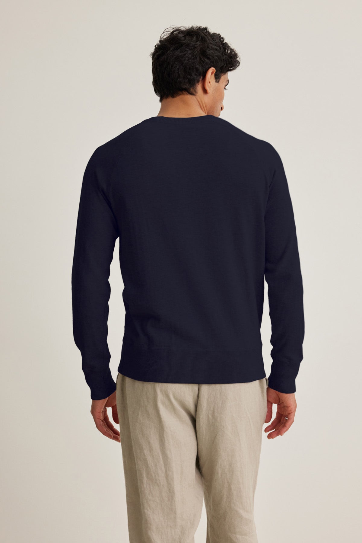 A man faces away from the camera, wearing the DARIO SWEATER by Velvet by Graham & Spencer—a mid-weight navy cotton cashmere crewneck—paired with light beige pants, set against a plain light background.