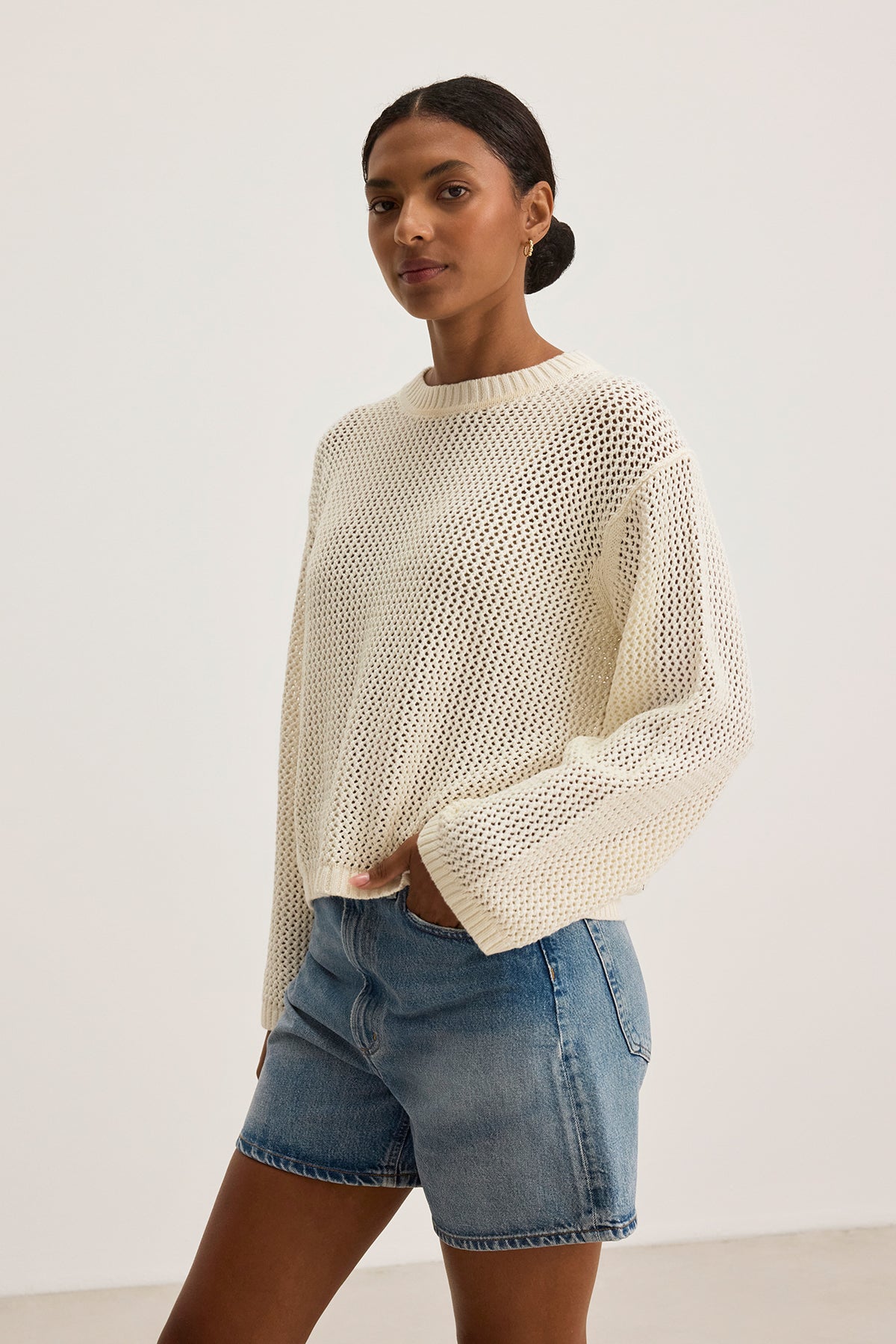   Against a plain backdrop, someone wears Velvet by Jenny Graham's KANAN COTTON CASHMERE MESH SWEATER with a crew neckline, paired with denim shorts. 