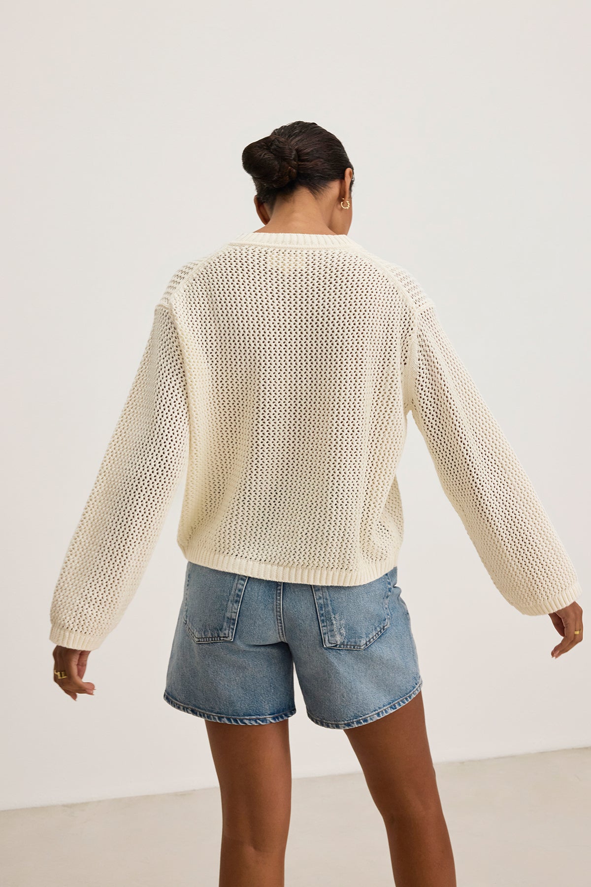   A person wearing the KANAN COTTON CASHMERE MESH SWEATER from Velvet by Jenny Graham and denim shorts stands with their back to the camera in a plain space. 