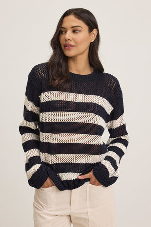 A woman models the Velvet by Jenny Graham KANAN COTTON CASHMERE MESH SWEATER, featuring black and white stripes with a crew neckline, paired with beige pants against a plain background.