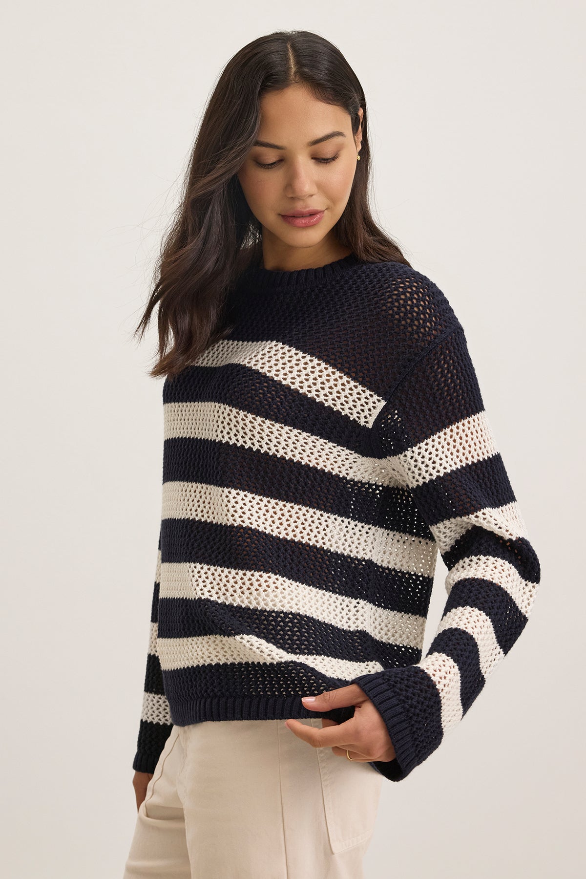   A person in a KANAN COTTON CASHMERE MESH SWEATER by Velvet by Jenny Graham with black and white stripes, crew neckline, and light trousers looks down to the side against a plain backdrop. 