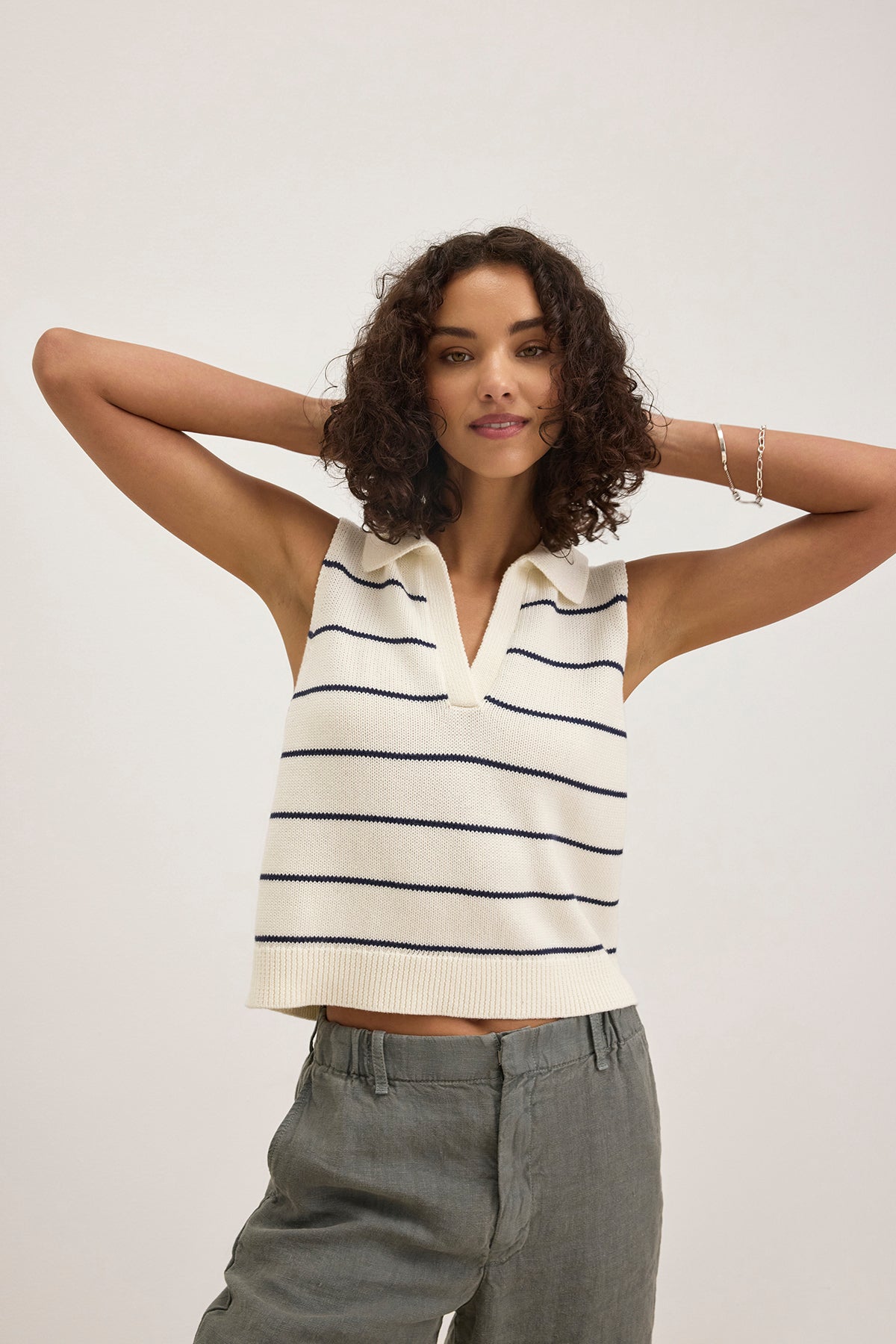 A woman wears the NAYLA COTTON CASHMERE SWEATER VEST by Velvet by Graham & Spencer—a sleeveless, collared white knit top with black stripes—paired with gray pants, standing with arms raised behind her head against a plain background.