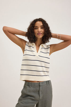 A woman wears the NAYLA COTTON CASHMERE SWEATER VEST by Velvet by Graham & Spencer—a sleeveless, collared white knit top with black stripes—paired with gray pants, standing with arms raised behind her head against a plain background.