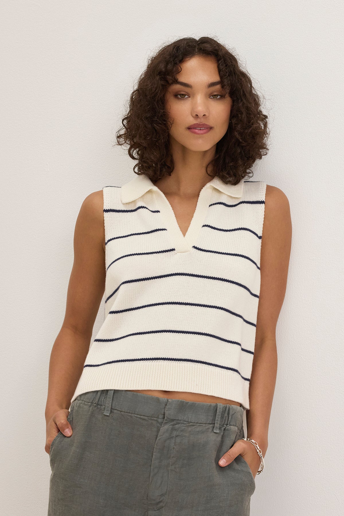 A woman stands against a white wall wearing the NAYLA COTTON CASHMERE SWEATER VEST by Velvet by Graham & Spencer—sleeveless, collared, with black horizontal stripes—paired with gray pants for a modern, layered look.