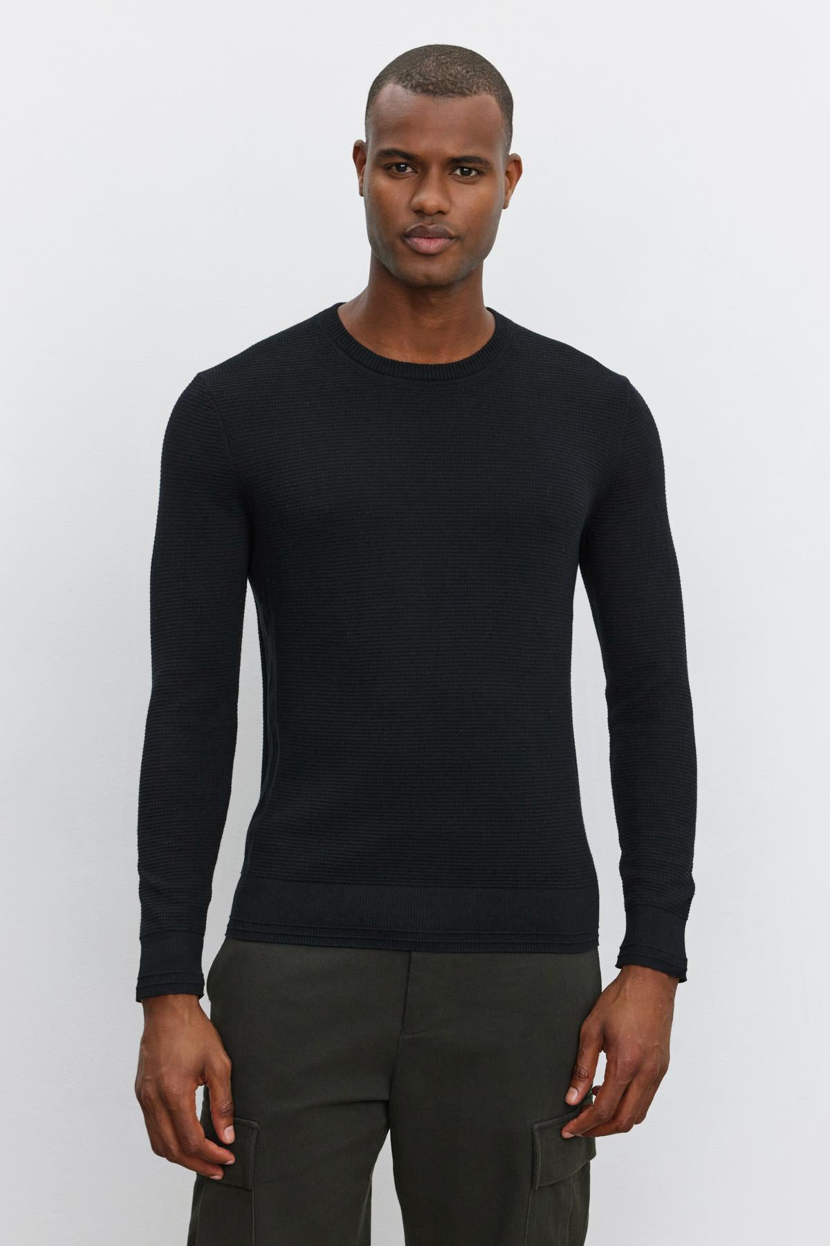   A man with a neutral expression wears the Velvet by Graham & Spencer WALTER SWEATER, a black long-sleeved top made of cotton and cashmere, featuring a crew neckline. He pairs it with dark green cargo pants while standing against a plain white background. 