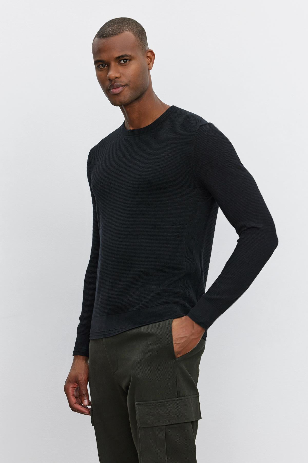   A person stands against a plain white background, wearing the WALTER SWEATER by Velvet by Graham & Spencer, a black long-sleeve shirt with a crew neckline, paired with green cargo pants. One hand is in their pocket while the other hangs by their side. 