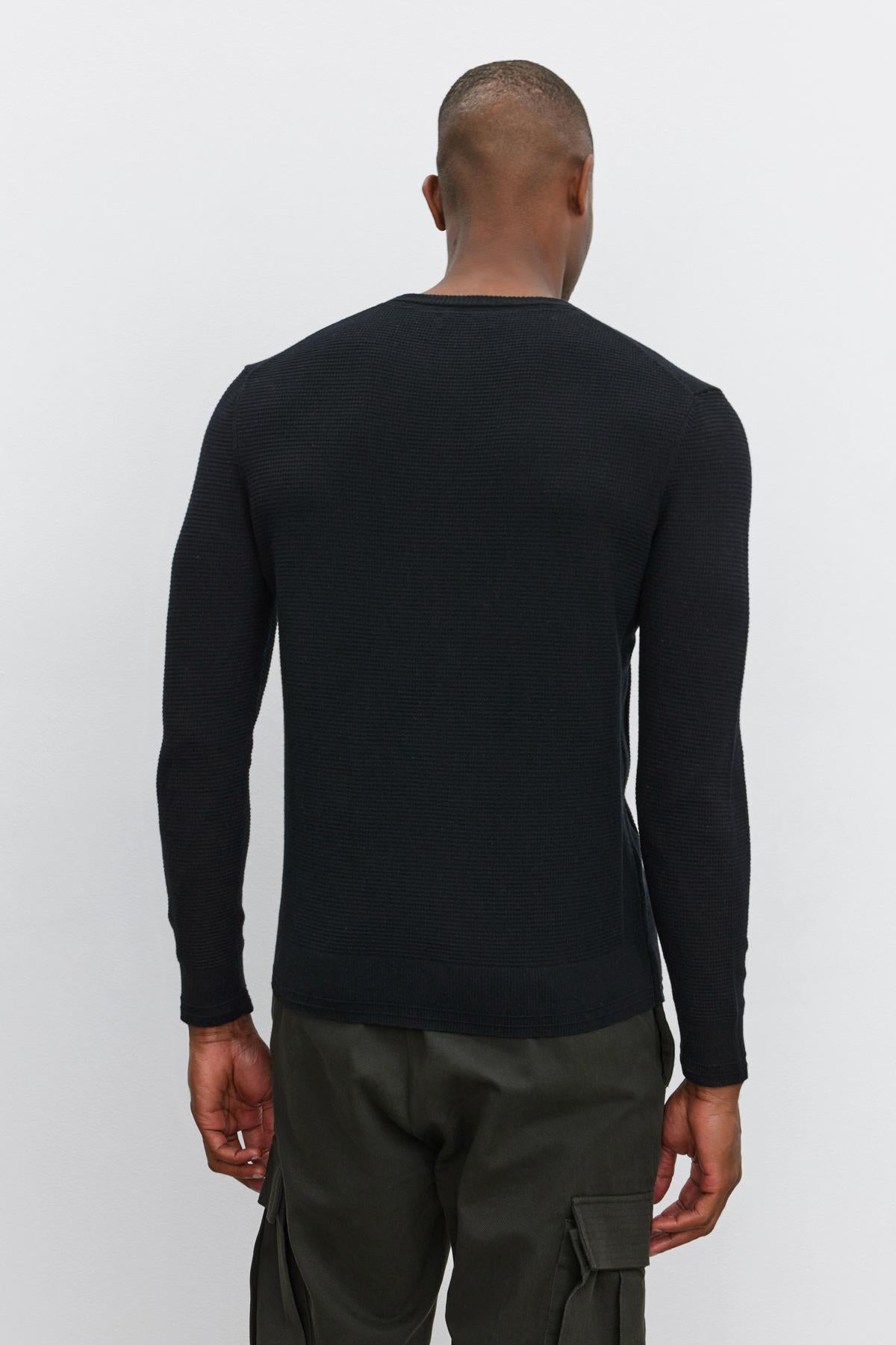   A person wearing a black WALTER SWEATER by Velvet by Graham & Spencer, featuring a classic crew neckline, paired with green cargo pants, facing away and standing against a plain white background. 