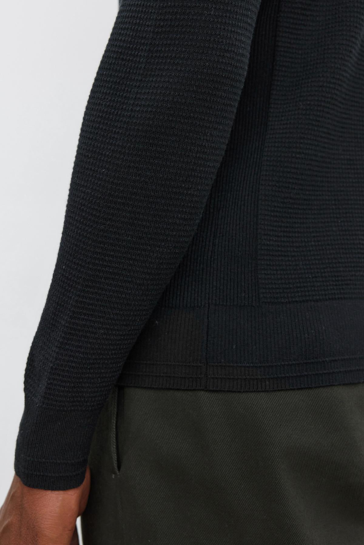   Close-up of a person wearing the WALTER SWEATER by Velvet by Graham & Spencer, featuring a textured black long-sleeve design with a crew neckline, paired with dark green pants and highlighting the arm and side of the torso. 