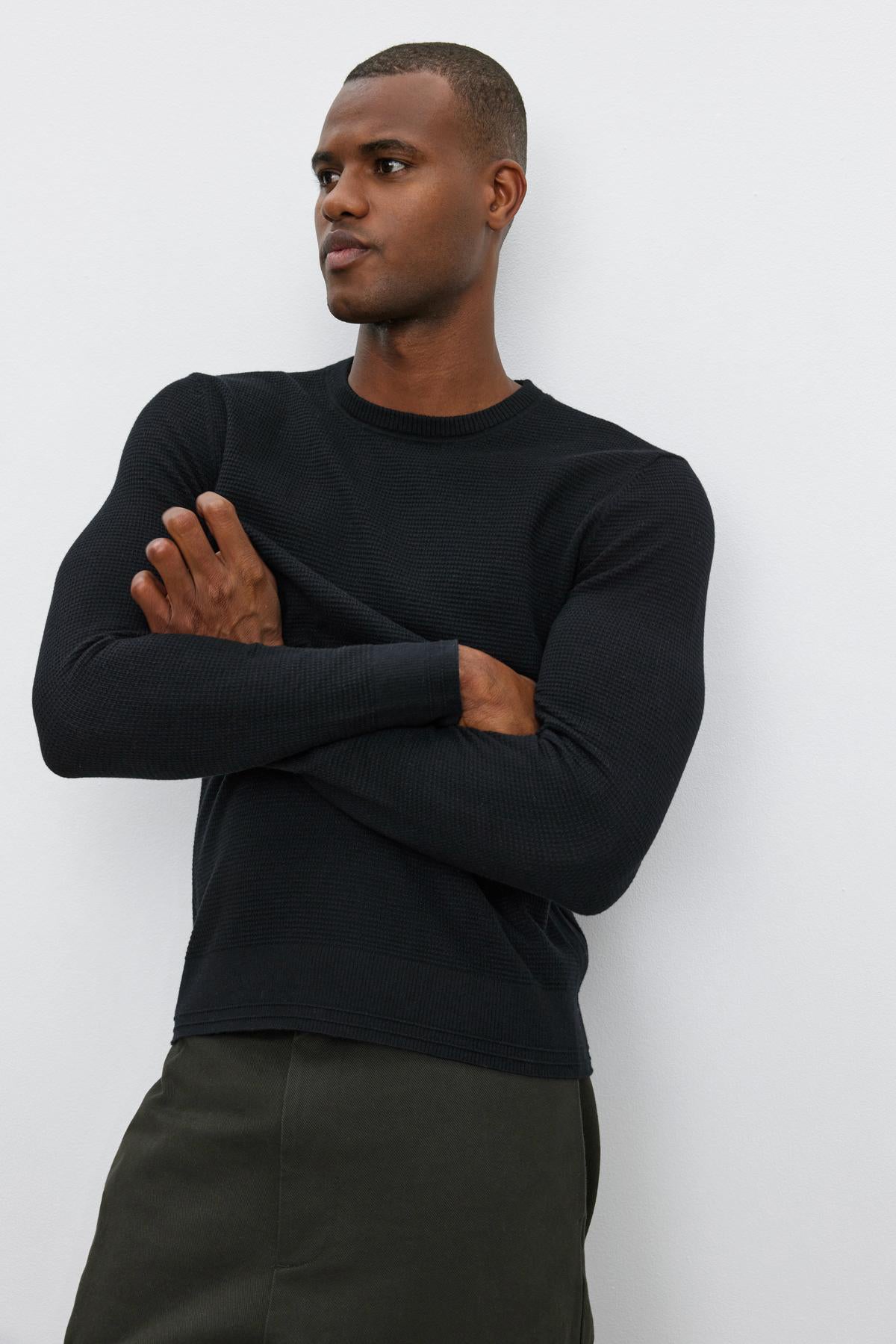   A person in a WALTER SWEATER by Velvet by Graham & Spencer, featuring long sleeves and a crew neckline, paired with dark pants, stands against a plain white background with arms crossed, looking to the side. 