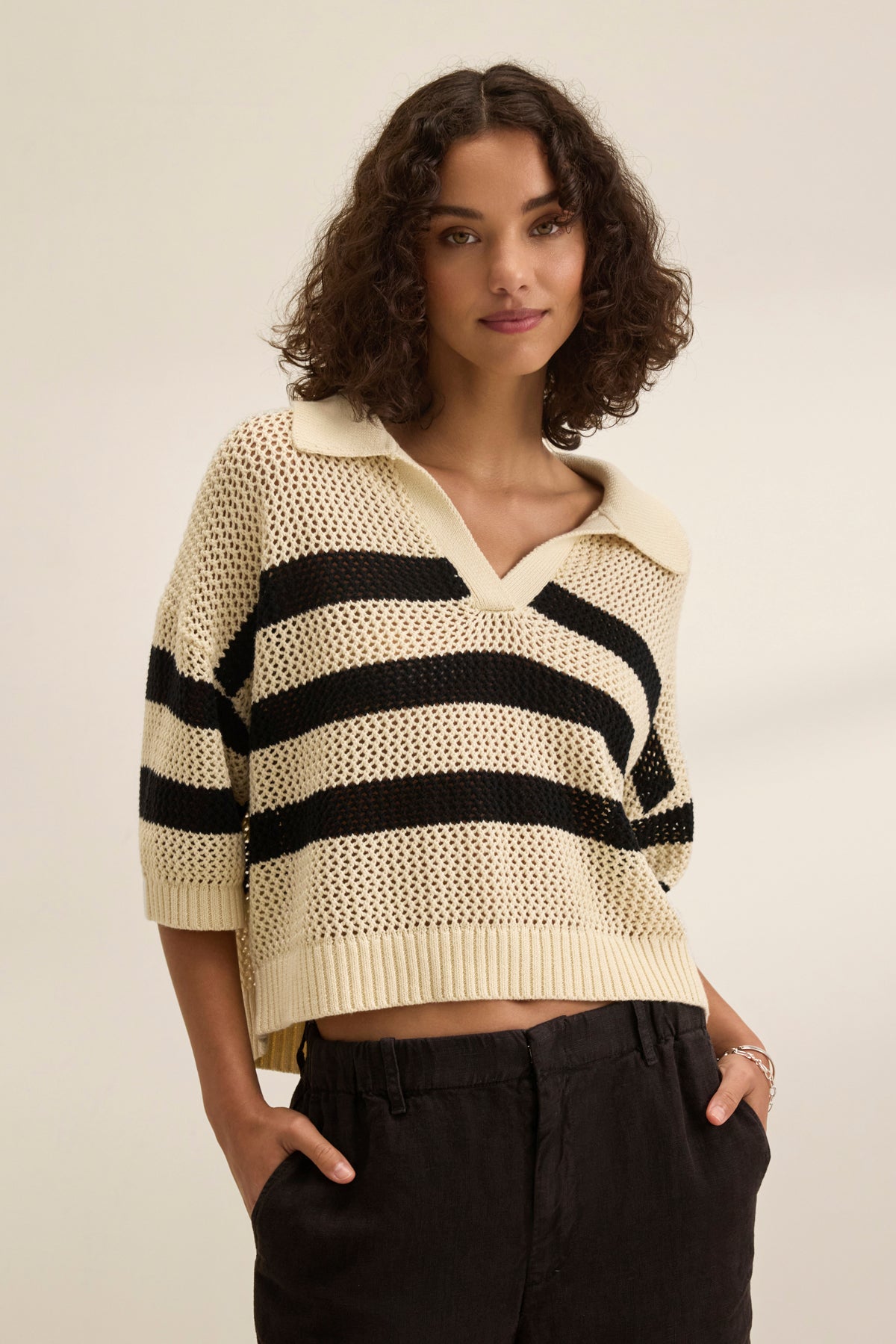 Wearing a striped YORK COTTON CASHMERE MESH SWEATER POLO by Velvet by Jenny Graham and dark pants, a person with curly hair exudes casual charm against a plain background.-38928816144577