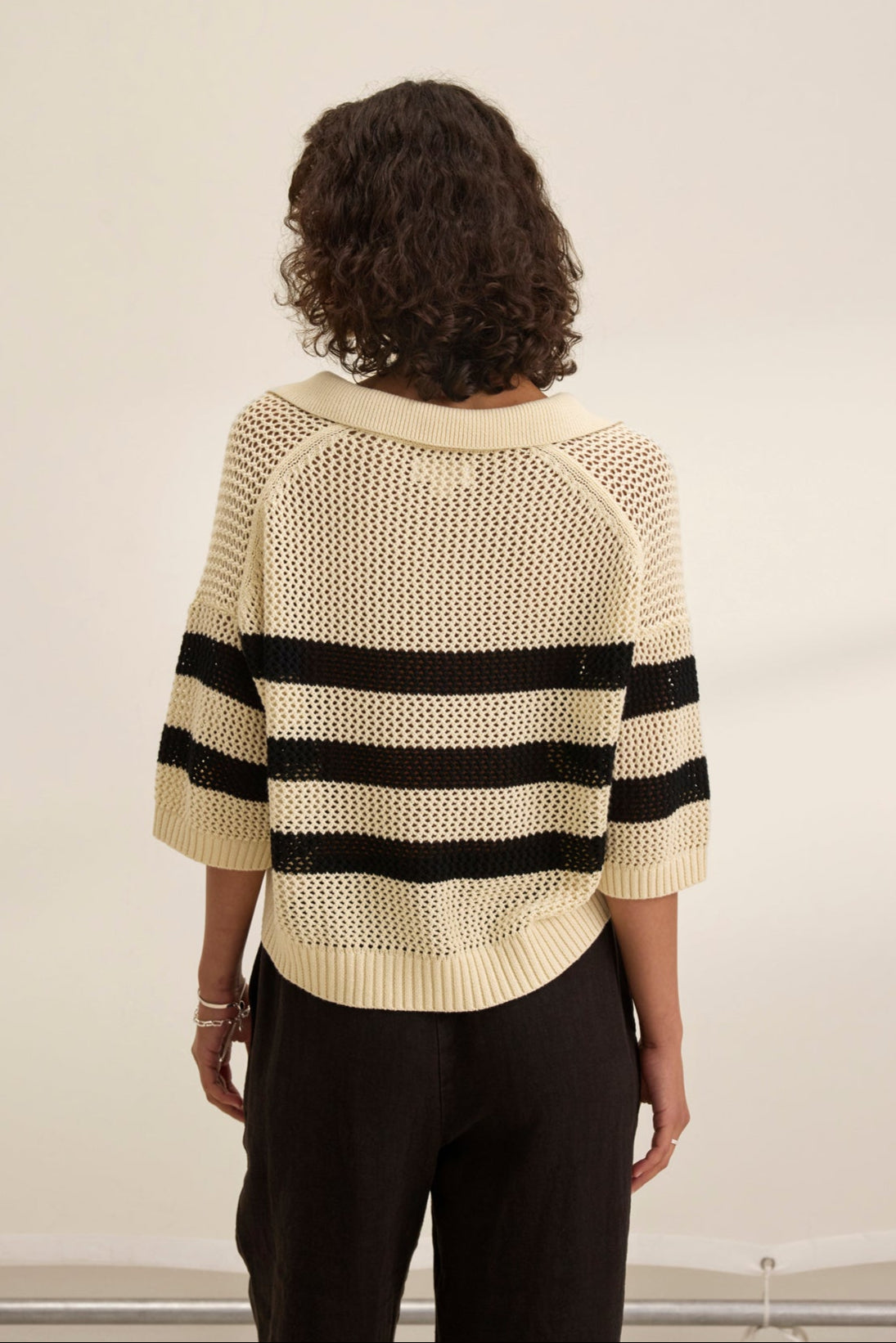 A person with curly hair faces away, wearing a Velvet by Jenny Graham YORK COTTON CASHMERE MESH SWEATER POLO in beige with black horizontal stripes, paired with black pants against a plain background.-38928816046273