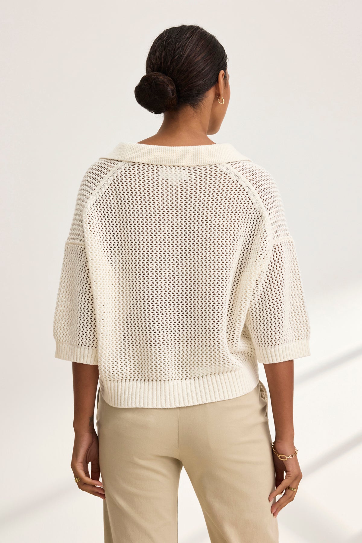 With hair in a bun, they stand facing away, dressed in a YORK COTTON CASHMERE MESH SWEATER POLO by Velvet by Jenny Graham. Their beige pants enhance the attire's soft elegance.-38928816275649