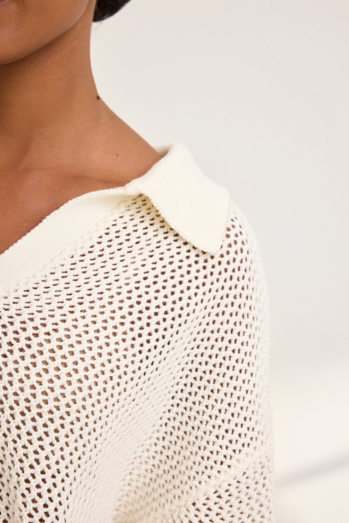 Close-up of a person wearing a YORK COTTON CASHMERE MESH SWEATER POLO by Velvet by Jenny Graham in white, exuding casual charm with its open-knit design that reveals the shoulder and part of the neck against a plain backdrop.-38928816242881