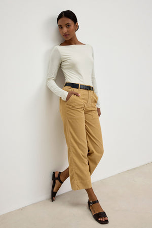 A woman stands against a white wall, wearing a long-sleeved white top, the VERA CORDUROY PANT by Velvet by Graham & Spencer—tan high-waisted cropped wide-leg pants with utility details—a black belt, and black sandals.