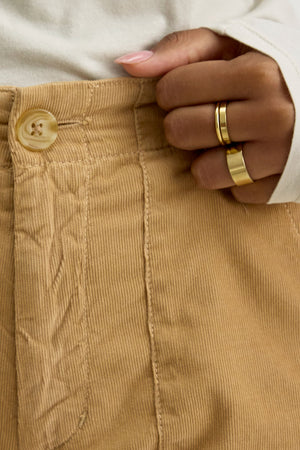 Close-up of a person wearing the VERA CORDUROY PANT by Velvet by Graham & Spencer in beige pinwale corduroy, featuring a button and top stitching, holding the waistband with a hand adorned with gold rings.