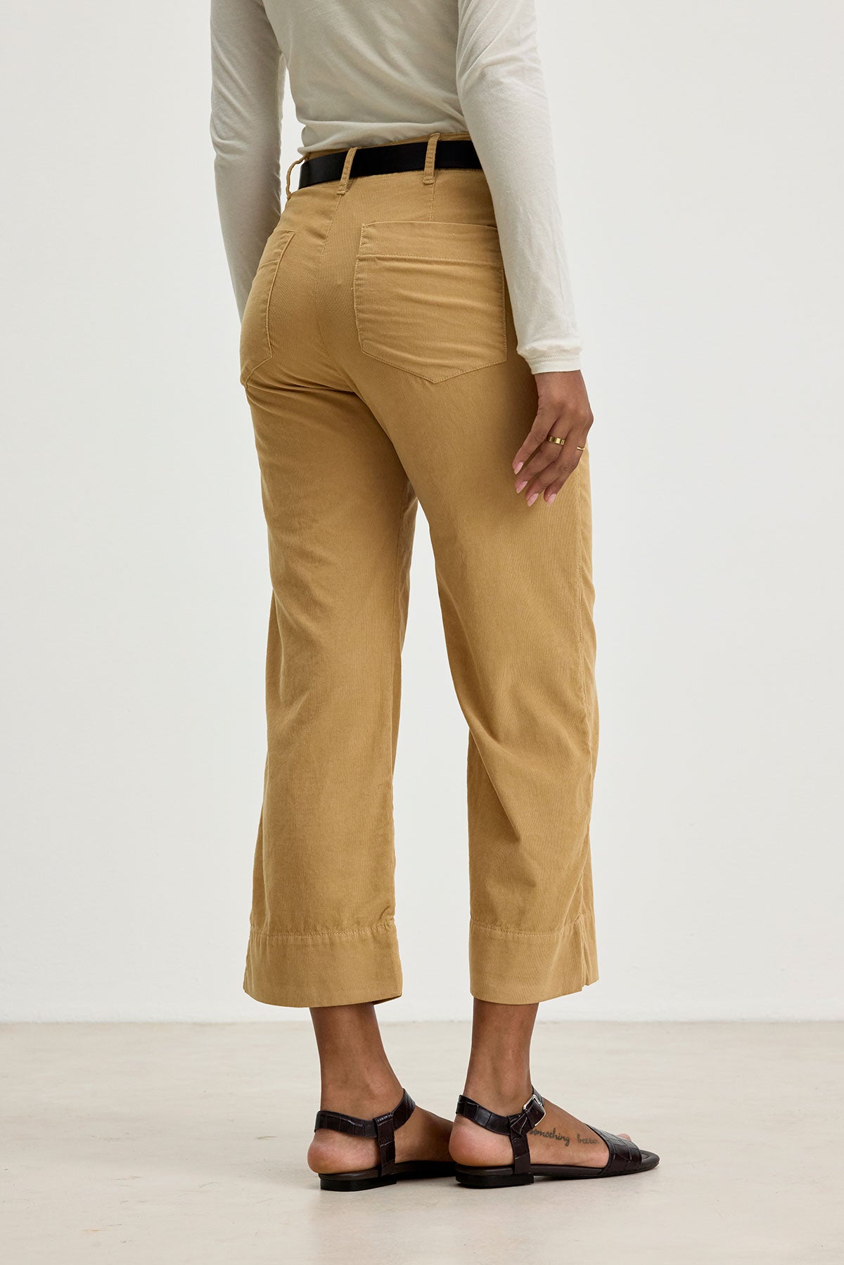 A person wearing the VERA CORDUROY PANT by Velvet by Graham & Spencer in beige, paired with a white long-sleeved top and sandals, is shown from the back. The person is standing in a well-lit, minimalist indoor setting.-37745668817089