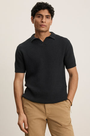 A man wears the Velvet by Graham & Spencer CARSTON SWEATER POLO, a modern fit textured black short-sleeve shirt, paired with khaki pants, standing against a plain light background.