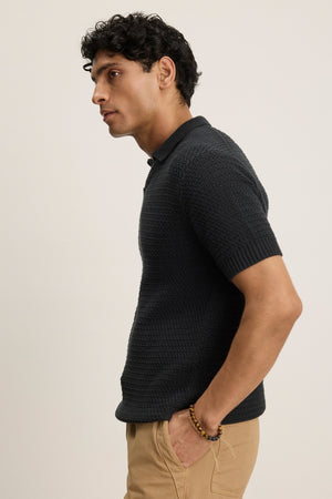 A man wearing the CARSTON SWEATER POLO by Velvet by Graham & Spencer and tan pants stands in profile with one hand in his pocket against a plain background.