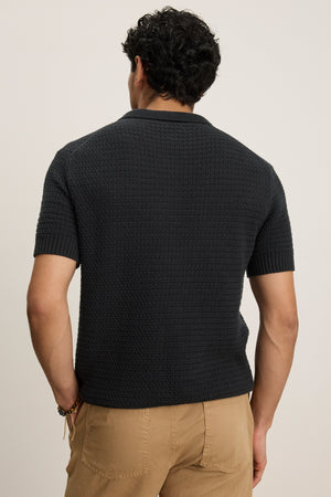 A man with short, curly hair is seen from the back wearing the CARSTON SWEATER POLO by Velvet by Graham & Spencer, a lightweight textured dark short-sleeve shirt, paired with light brown pants against a plain background.