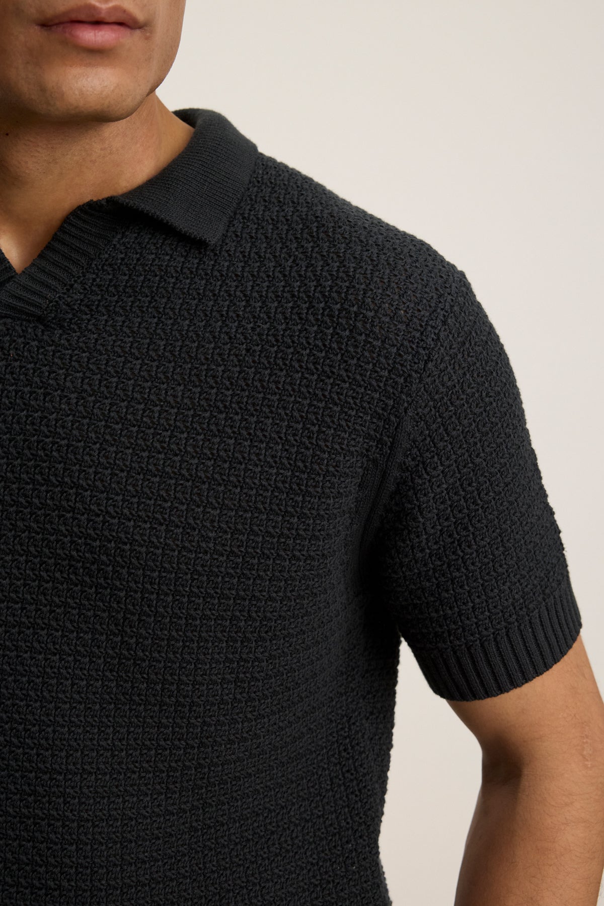 A close-up of a person wearing the CARSTON SWEATER POLO by Velvet by Graham & Spencer—a breathable, textured, short-sleeve black knit polo with a modern fit—against a plain background.-39446023635137