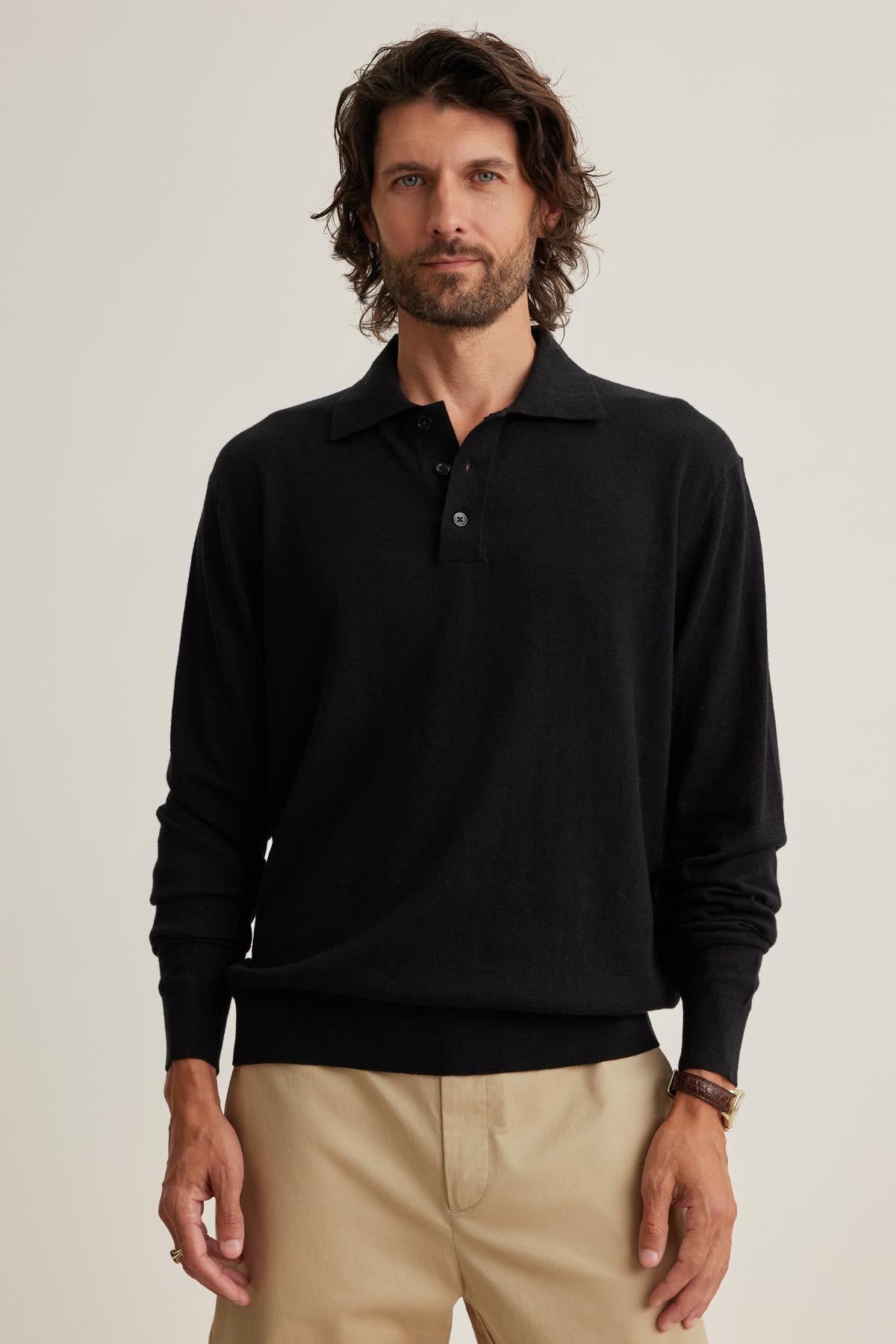   A man with wavy hair is wearing a black Velvet by Graham & Spencer MATTEO Cotton Linen Sweater Polo and beige pants while standing against a plain background. 