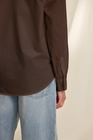 A person is seen from the back wearing the KELIA SHIRT by Velvet by Graham & Spencer in brown with light blue jeans, one arm visible and hand relaxed at their side.