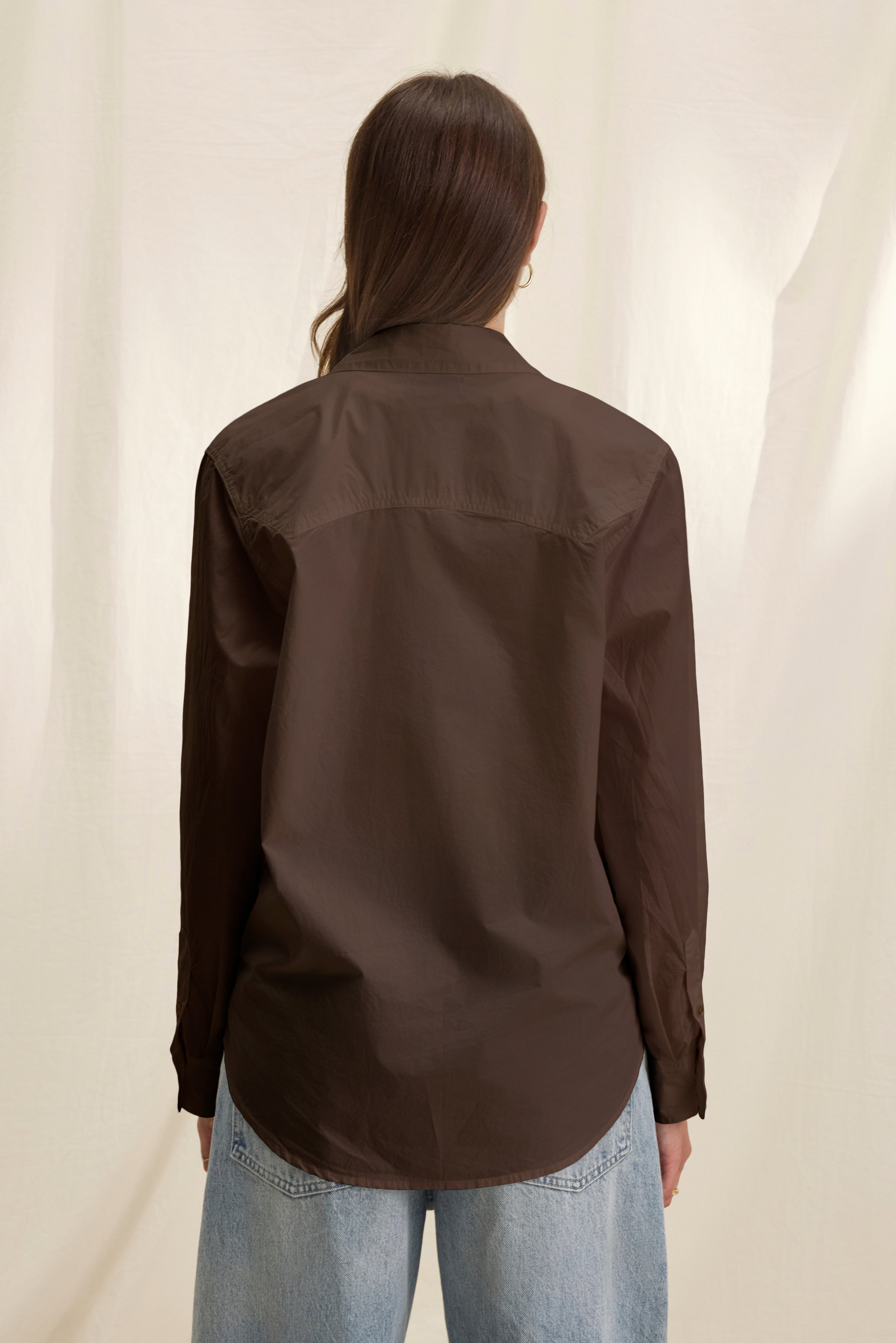 A person with long brown hair, wearing the KELIA SHIRT by Velvet by Graham & Spencer in dark brown and light blue jeans, stands facing away from the camera against a neutral background.