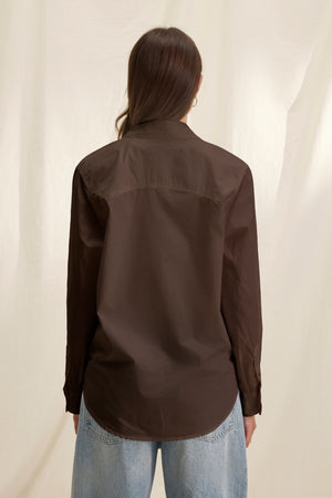 A person with long brown hair, wearing the KELIA SHIRT by Velvet by Graham & Spencer in dark brown and light blue jeans, stands facing away from the camera against a neutral background.