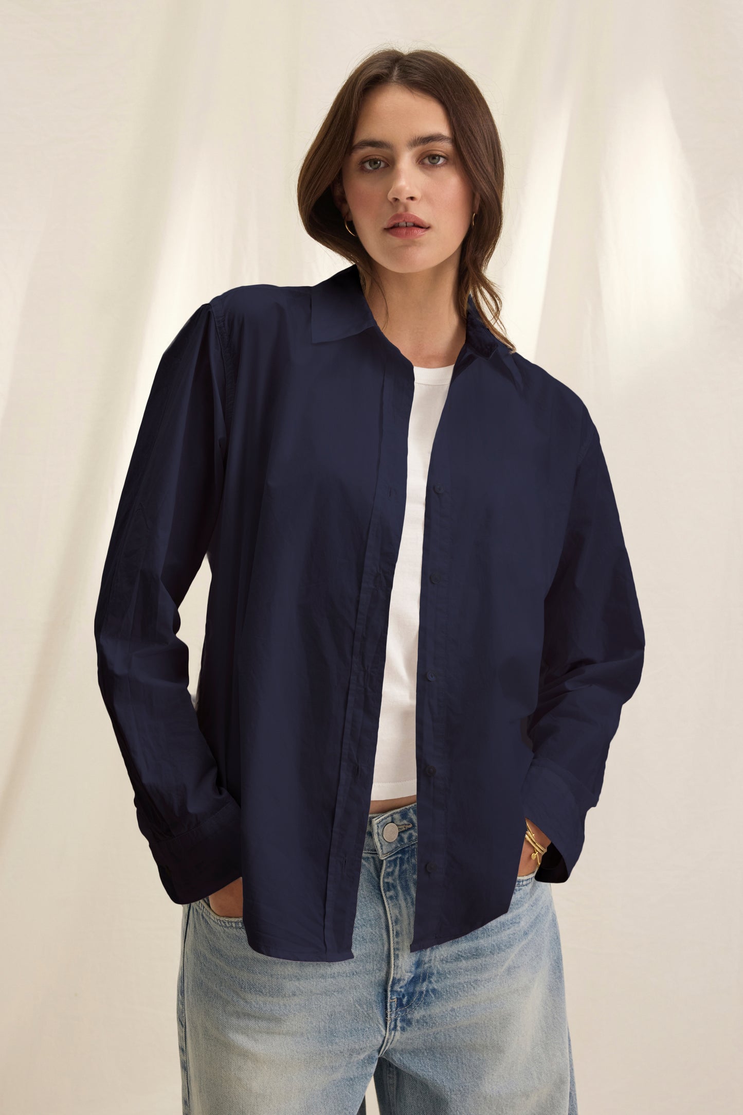 A woman stands in front of a neutral fabric backdrop wearing the Velvet by Graham & Spencer KELIA SHIRT, an oversized button-up in navy cotton poplin, layered over a white t-shirt and faded blue jeans.-40281878364353