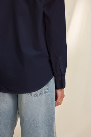Model wearing the KELIA SHIRT by Velvet by Graham & Spencer—a navy blue cotton poplin oversized button-up—paired with light blue jeans, shown from the back and side against a beige background.