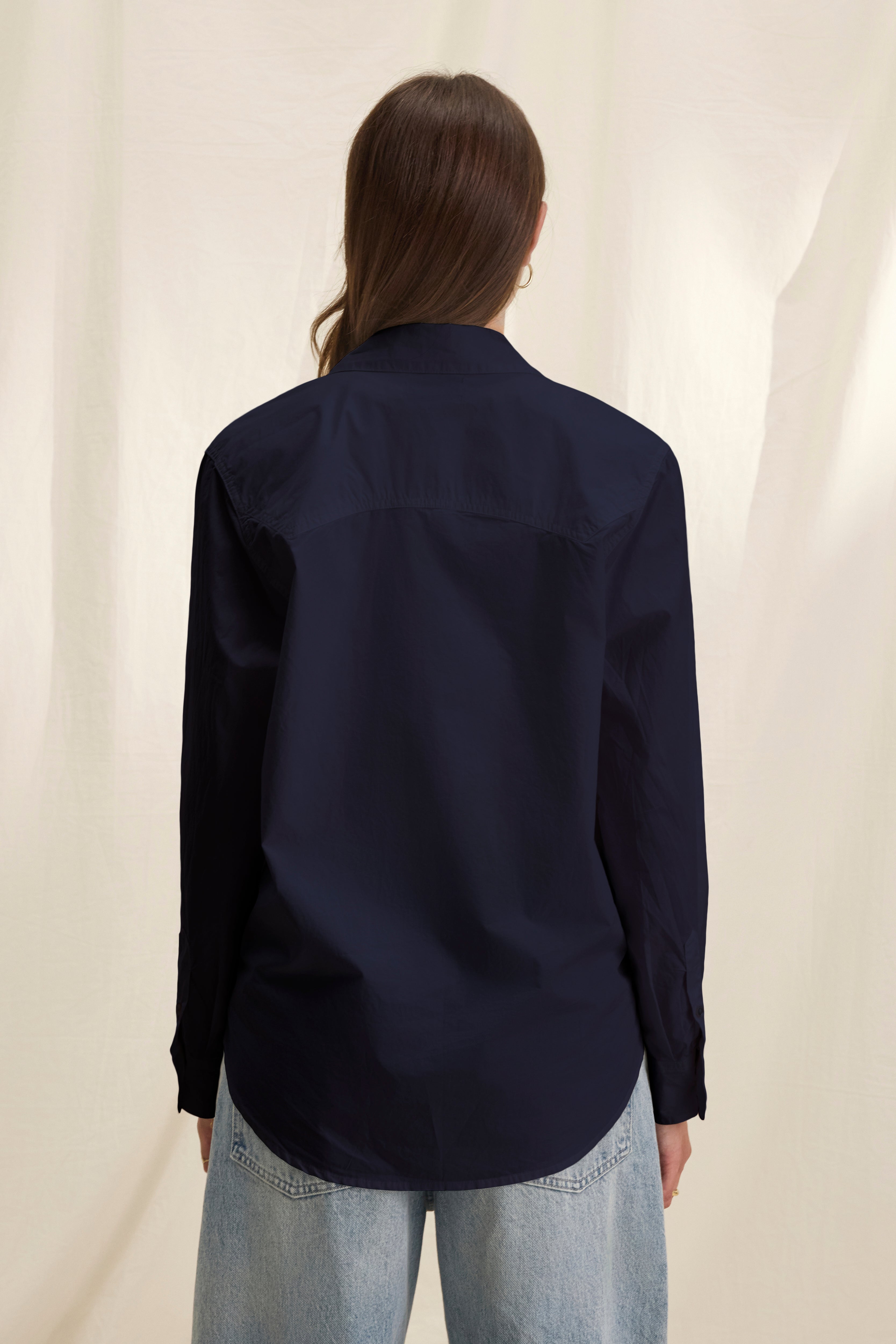 A person with long brown hair wears the KELIA SHIRT in dark navy cotton poplin by Velvet by Graham & Spencer and light blue jeans, standing with their back to the camera against a neutral background for a comfortable look.