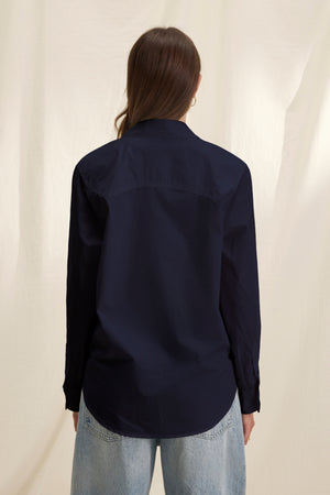 A person with long brown hair wears the KELIA SHIRT in dark navy cotton poplin by Velvet by Graham & Spencer and light blue jeans, standing with their back to the camera against a neutral background for a comfortable look.