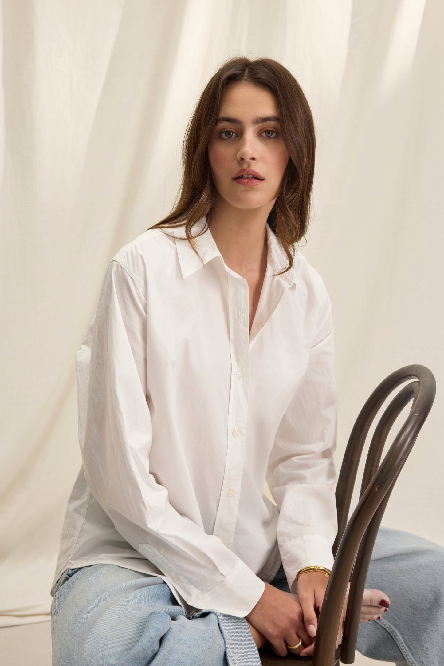 A woman with long brown hair wears the KELIA SHIRT by Velvet by Graham & Spencer and light blue jeans, seated on a wooden chair against a light fabric backdrop.-40281806078145
