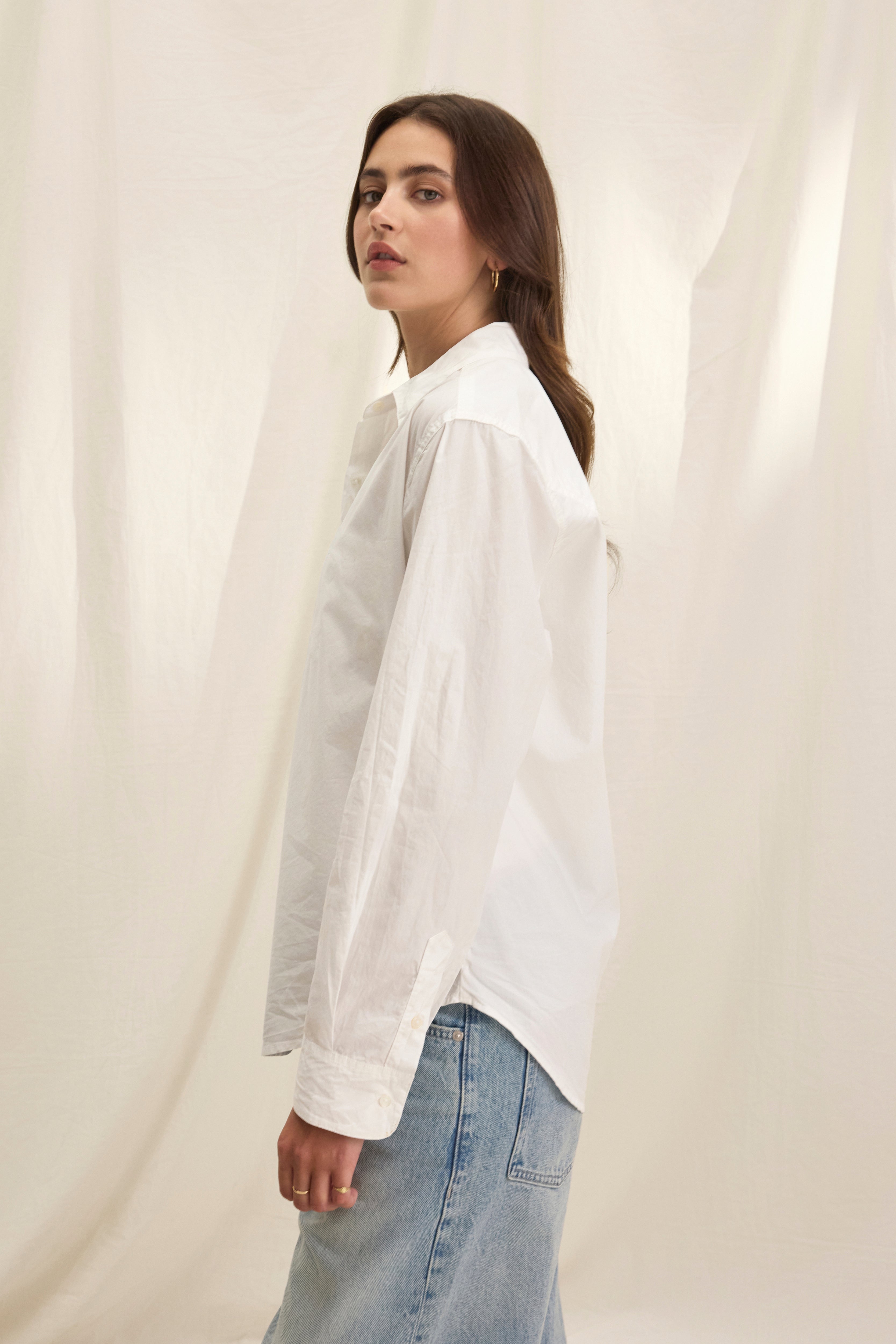A person in the KELIA SHIRT by Velvet by Graham & Spencer and light blue jeans stands sideways before a light fabric backdrop.